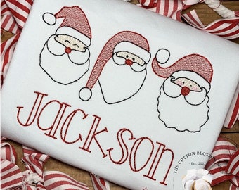 May include: White embroidered Christmas shirt with three red Santa faces and the name "Jackson" in red thread.