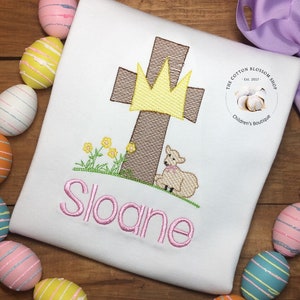 May include: White embroidered shirt featuring a brown cross, a yellow crown, a white lamb, and green grass. The name "Sloane" is embroidered in pink below the image. The shirt has a small white circle with the text "The Cotton Blossom Shop Est. 2017" and "Children's Boutique" in black text.