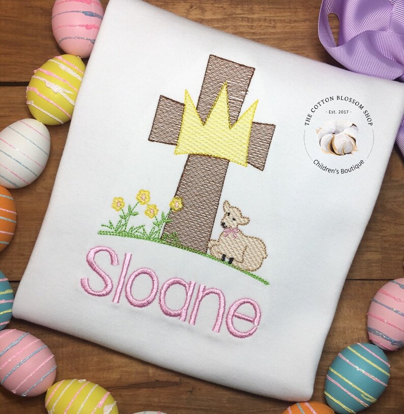May include: White embroidered shirt featuring a brown cross, a yellow crown, a white lamb, and green grass. The name "Sloane" is embroidered in pink below the image. The shirt has a small white circle with the text "The Cotton Blossom Shop Est. 2017" and "Children's Boutique" in black text.