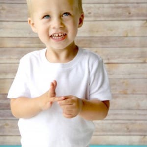 May include: A young boy with a spiky blond hairstyle is wearing a white t-shirt and smiling at the camera.