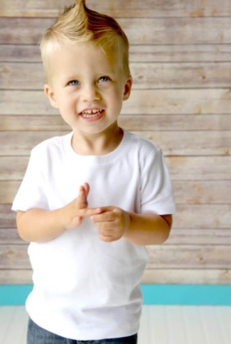 May include: A young boy with a spiky blond hairstyle is wearing a white t-shirt and smiling at the camera.