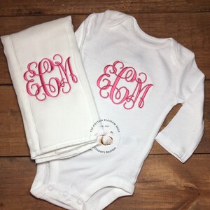 Puede incluir: Un body blanco para bebé y una manta blanca para recibir con un monograma bordado en rosa "EGM". El body tiene mangas largas y cierre a presión. La manta está doblada por la mitad. El texto "The Cotton Blossom Shop Est. 2017 Children's Boutique" está impreso en el body.
