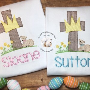 May include: Two white t-shirts with embroidered images of a brown cross with a yellow crown, a white lamb, and green grass. The text "Sloane" is embroidered in pink on one shirt and "Sutton" is embroidered in blue on the other shirt.