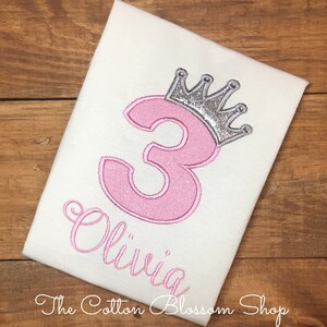 Puede incluir: Una camiseta blanca con un número 3 rosa y una corona plateada con purpurina en la parte superior. El nombre "Olivia" está escrito en cursiva rosa debajo del número. La camiseta está sobre una superficie de madera.