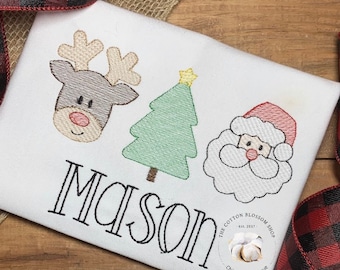 May include: White Christmas-themed embroidered shirt with a reindeer, Christmas tree, and Santa Claus design. The name "Mason" is embroidered below the design. The shirt has a small logo for "The Cotton Blossom Shop" in the bottom right corner.