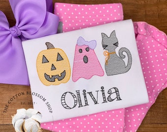 May include: White t-shirt with embroidered Halloween designs: a jack-o'-lantern, a ghost, and a cat. The name "Olivia" is embroidered below the designs. A large purple bow and pink polka dot fabric are in the background.