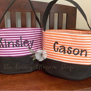 May include: Two striped fabric trick-or-treat bags with black handles. The bag on the left is purple and white with the name "Kinsley" embroidered on it. The bag on the right is orange and white with the name "Gason" embroidered on it. Both bags have a black bottom.