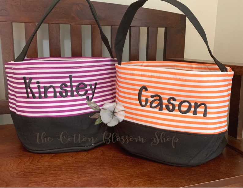May include: Two striped fabric trick-or-treat bags with black handles. The bag on the left is purple and white with the name "Kinsley" embroidered on it. The bag on the right is orange and white with the name "Gason" embroidered on it. Both bags have a black bottom.