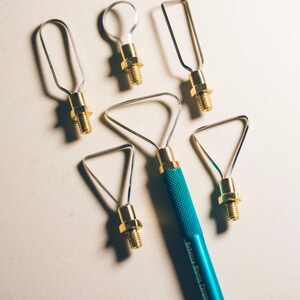 May include: A collection of ceramic tools with metal loops and triangular shapes, connected to brass fittings and teal handles. One handle is inscribed with "Rebecca Morris Ceramics". These tools are designed for shaping and sculpting clay.