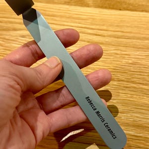 Peut inclure: Un outil en métal argenté avec une extrémité carrée et un manche arrondi. Le manche est gravé du texte "Rebecca Morris Ceramics".