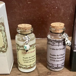 Peut inclure: Deux petits flacons en verre avec des bouchons en liège et des breloques décoratives, ainsi que deux boîtes rectangulaires. Les flacons et les boîtes présentent des étiquettes avec du texte français, dont "Le labo de Cléopâtre" et "Poudre de Chypre".