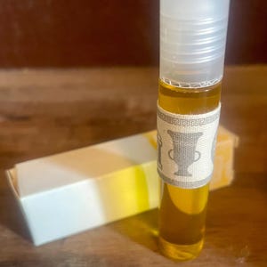 Könnte beinhalten: Eine klare Glasflasche mit goldener Flüssigkeit. Die Flasche ist mit einem weißen Etikett mit einem grauen Design einer Vase und einem geometrischen Muster umwickelt. Die Flasche steht auf einer Holzoberfläche.