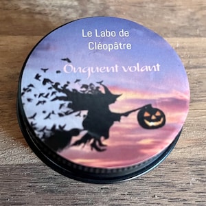 May include: A round, black tin container with a purple and pink sunset scene. The tin has a black and white silhouette of a witch flying on a broomstick with a jack-o-lantern. The text on the tin reads "Le Labo de Cléopâtre Onguent volant".