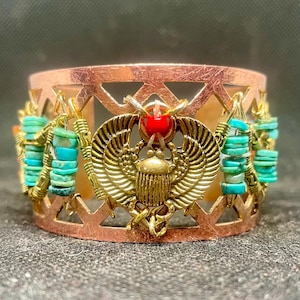 Può includere: Bracciale a polsino color rame con un design geometrico ritagliato. Il bracciale presenta uno scarabeo dorato con dettagli rossi e perline turchesi. Il bracciale è una fascia larga, che mostra un tema egiziano.