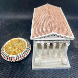 Può includere: Un modello in miniatura di un tempio classico con colonne bianche e un tetto di tegole rosse. Accanto, un piccolo piatto ovale con un bordo decorativo rosso e bianco, riempito con una sostanza strutturata di colore giallo.