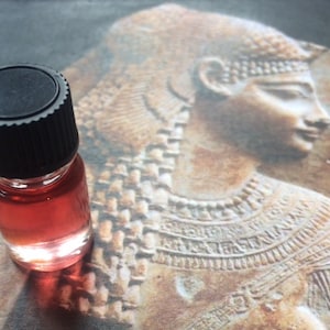 May include: A small, clear glass bottle with a black screw-top lid. The bottle is filled with a red liquid. The bottle is sitting on a brown, textured surface with a carved image of a woman's head.