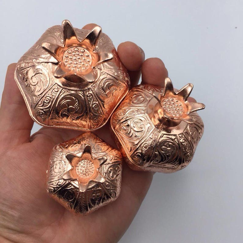 May include: Three copper pomegranate shaped boxes with intricate designs. The boxes are different sizes and are arranged in a hand.