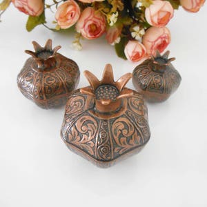 May include: Three small decorative boxes shaped like pomegranates. The boxes are made of copper and have an intricate floral design. The boxes are a dark brown color with copper accents.