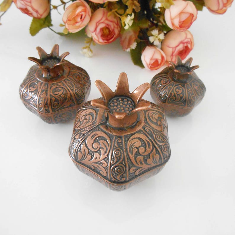 May include: Three small decorative boxes shaped like pomegranates. The boxes are made of copper and have an intricate floral design. The boxes are a dark brown color with copper accents.