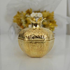 May include: A gold-colored metal pomegranate-shaped box with intricate embossed designs. The box has a hinged lid and a decorative crown-like top.