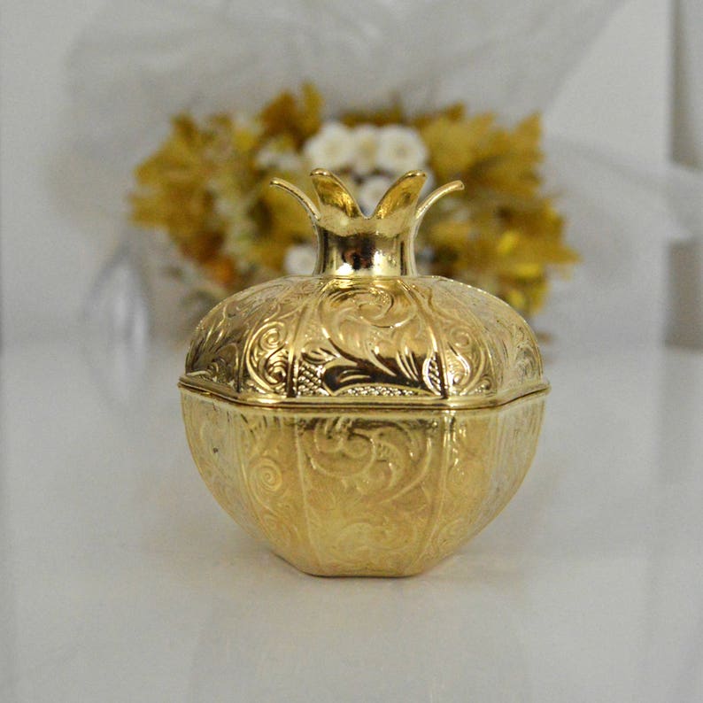 May include: A gold-colored metal pomegranate-shaped box with intricate embossed designs. The box has a hinged lid and a decorative crown-like top.