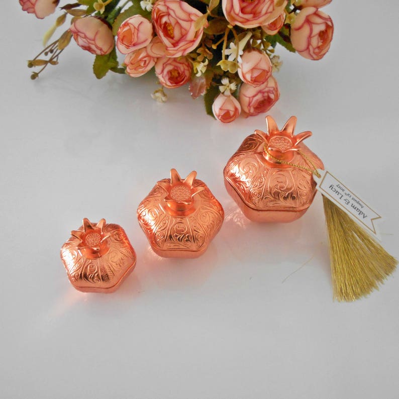 May include: Three copper-colored pomegranate-shaped boxes with intricate designs. The boxes are arranged in a row from largest to smallest. The largest box has a tag attached that says "Happy New Year!" in gold lettering.