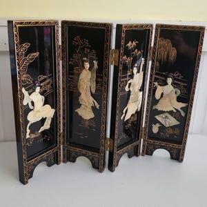 May include: A four-panel folding screen with a glossy black finish. Each panel features a different scene of Asian figures and landscapes, with gold accents and mother-of-pearl inlays. The screen is hinged with brass hardware and has decorative feet.