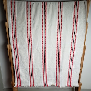 May include: A cream-colored textile with vertical stripes of red, black, and white. The fabric is displayed on a wooden drying rack. The stripes are evenly spaced and run the length of the textile, creating a simple, yet striking pattern.