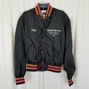 Può includere: Giacca nera in nylon con finiture a righe rosse e arancioni su colletto, polsini e vita. La giacca ha chiusure a scatto rosse e due tasche anteriori. Il nome "Andy" è ricamato sul petto sinistro e il testo sul petto destro recita "Portland Plastic Pipe, Portland Babe Ruth League Champion 1991."
