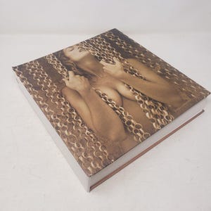 May include: A book with a white cover and a brown spine. The cover features a sepia-toned photograph of a woman with long hair, posing with chains.