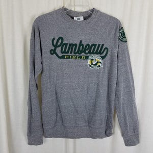 May include: Gray sweatshirt with the words "Lambeau Field" in green script and a green and yellow logo. The sweatshirt has long sleeves and a crew neck. A circular patch with text is on the right sleeve.