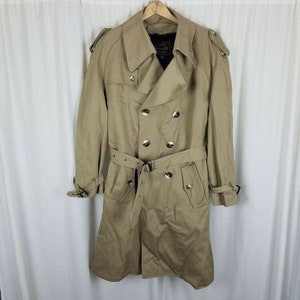 Könnte beinhalten: Ein beigefarbener Trenchcoat mit doppelreihigem Design, silbernen Knöpfen und passendem Gürtel. Der Mantel hat Schulterklappen und hängt an einem Holzbügel. Ein klassisches Oberbekleidungsstück.