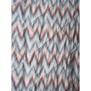 May include: Close-up of a quilted fabric with a repeating chevron pattern. The design features alternating rows of blue, peach, and white zigzags, creating a textured appearance. Diagonal stitching adds a quilted effect.