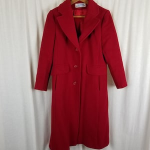 May include: A long, red wool coat with a classic design. The coat features a notched collar, two front pockets, and a three-button closure. The coat is hanging on a wooden hanger.
