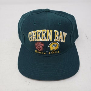 Può includere: Un cappellino da baseball verde scuro con visiera piatta. Il cappellino presenta la scritta "GREEN BAY" in oro e bianco. Sotto, due loghi ricamati e la scritta "Since 1921".