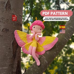 May include: A crocheted rosy maple moth amigurumi doll with pink and yellow wings, a pink head covering, and a yellow dress. The doll is attached to a tree branch. The image includes the text "PDF PATTERN".
