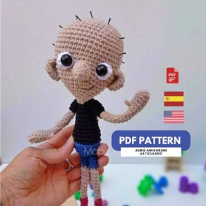Puede incluir: Muñeco amigurumi de crochet de un personaje de dibujos animados con piel marrón, cabello negro, una camiseta negra, pantalones cortos azules y zapatos rojos. El muñeco está levantando su brazo derecho en señal de saludo. El muñeco está etiquetado como "SUMO AMIGURUMI ARTICULADO" y "PDF PATTERN".