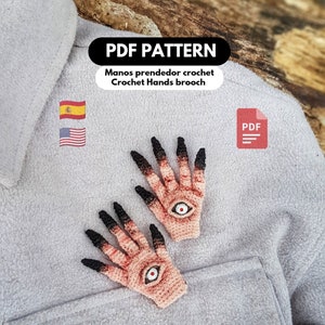 Puede incluir: Patrón PDF para tejer al crochet un par de manos con ojos. Las manos son de color rosa claro con uñas negras y un ojo blanco en la palma de cada mano. El patrón está disponible en español e inglés. La imagen también incluye el texto "PDF PATTERN" y "Crochet Hands brooch".