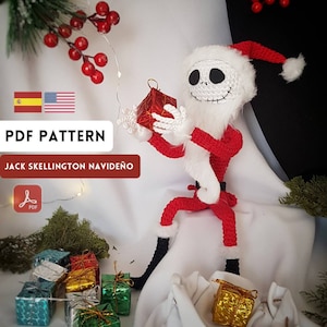 Puede incluir: Patrón de crochet para un adorno navideño de Jack Skellington. El patrón está disponible para descargar en PDF. La imagen muestra un Jack Skellington de crochet vestido de Papá Noel, sosteniendo una caja de regalo roja. Hay varios regalos envueltos en el fondo.