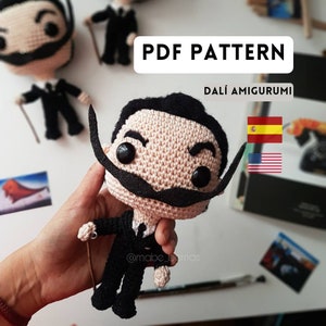 May include: A crocheted amigurumi doll of Salvador Dalí, a famous Spanish artist, wearing a black suit and a mustache. The doll is holding a cane in its hand. The image includes the text "PDF PATTERN DALÍ AMIGURUMI" and flags of Spain and the United States.