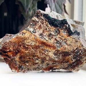 May include: A rough-hewn rock with a layered appearance. The rock features bands of brown, orange, and gray hues, with patches of green and black. The texture appears coarse and natural, suggesting a mineral specimen or decorative stone.