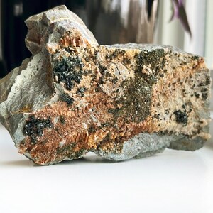 May include: A rough-hewn rock specimen with a layered appearance. The stone features a mix of gray, tan, and reddish-brown hues, with small dark mineral deposits scattered across its surface. The texture appears coarse and natural.