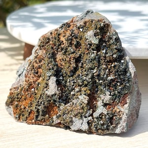 May include: A rough-hewn rock with a textured surface. The rock displays a mix of colors, including shades of brown, gray, and green. Small, dark crystals are scattered across the surface, adding a sparkling effect. The rock is a natural mineral specimen.