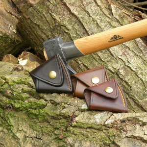 May include: Three leather axe sheaths in brown, dark brown, and black with a brass button closure. The sheaths are lying on a tree trunk with a wooden-handled axe in the background.