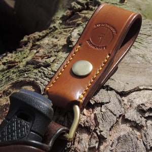 May include: A brown leather belt loop with a brass button and stitching. The loop has the text "STEALTHCRAFT" embossed on it.