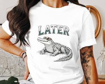 May include: White t-shirt featuring a detailed illustration of an alligator wearing sunglasses. The word "LATER" is printed above the alligator in a green and silver font. The design is centered on the shirt.