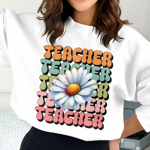 Puede incluir: Sudadera blanca con la palabra "TEACHER" en naranja, turquesa, verde y rosa, apilada verticalmente. Una gran margarita con pétalos blancos y centro amarillo está en el diseño. La sudadera es de estilo casual.