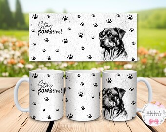 May include: A white ceramic mug featuring a black and white illustration of a Rottweiler dog, a pattern of black paw prints, and the text "Stay pawsitive!" printed in a black script font. The design wraps around the exterior of the mug.