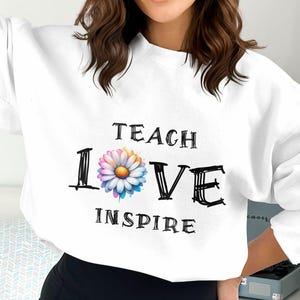 Puede incluir: Sudadera blanca con las palabras "TEACH LOVE INSPIRE" en letras negras. La palabra "LOVE" presenta una margarita colorida en lugar de la letra "O". Una parte superior informal y cómoda.