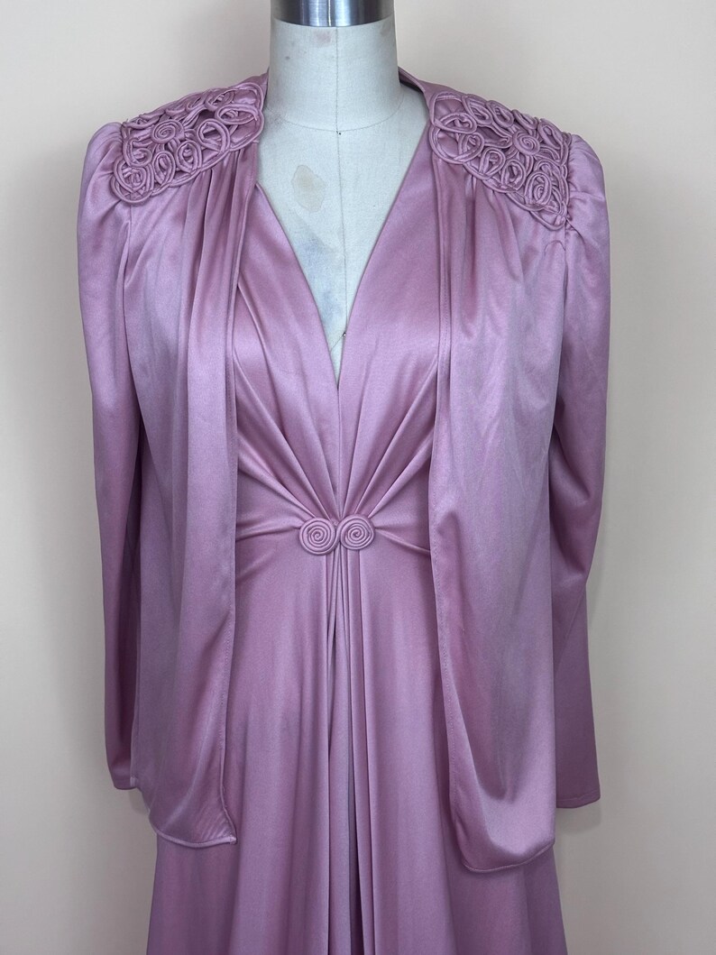 May include: A vintage lilac dress and jacket ensemble. The dress has a V-neck and a gathered waist with a decorative detail. The jacket features decorative shoulder accents and long sleeves. The outfit is displayed on a mannequin.